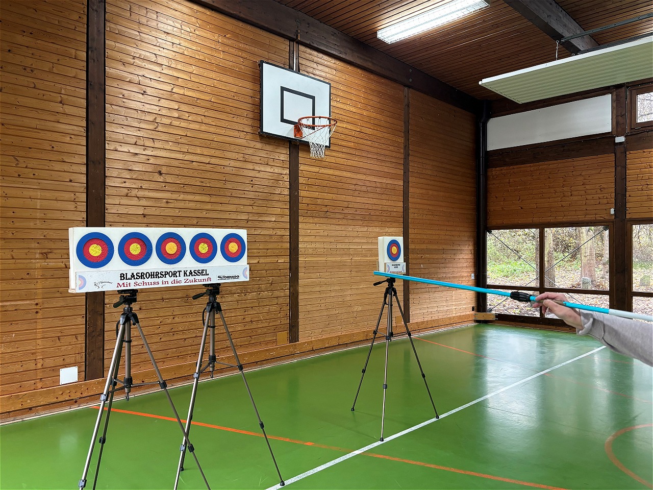 Blasrohrsport in einer Turnhalle. Zielscheiben stehen auf Stativen, eine Person zielt mit einem Blasrohr