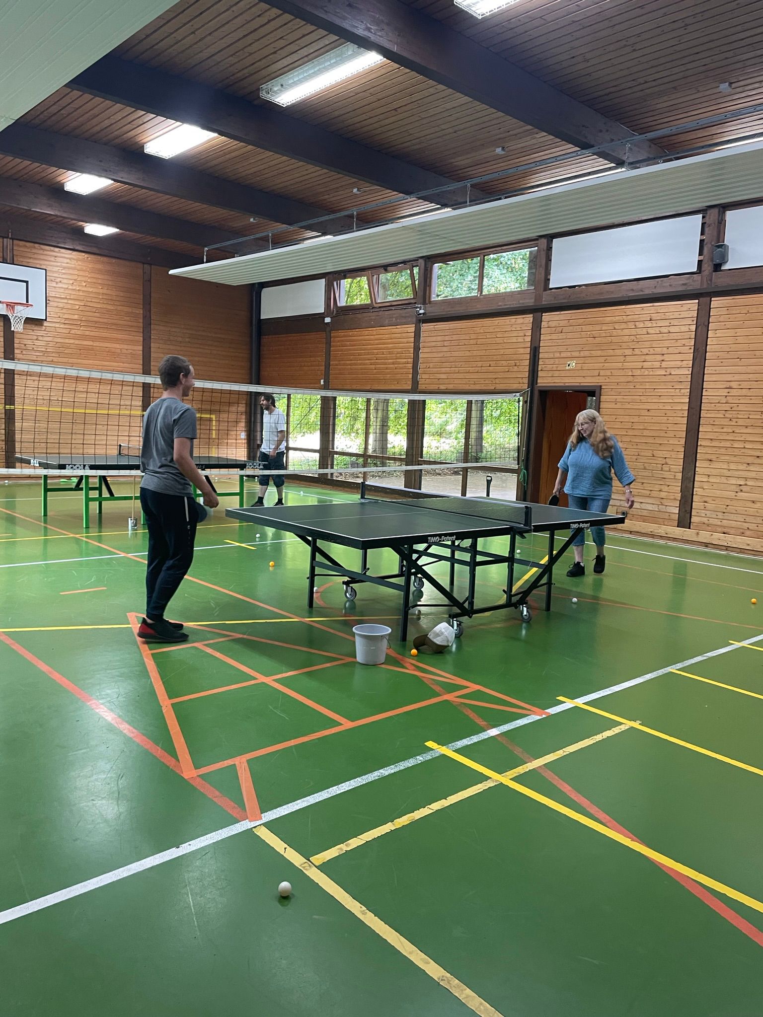 Zwei Personen spielen Tischtennis in einer Sporthalle mit Holzverkleidung und grünen Linien am Boden