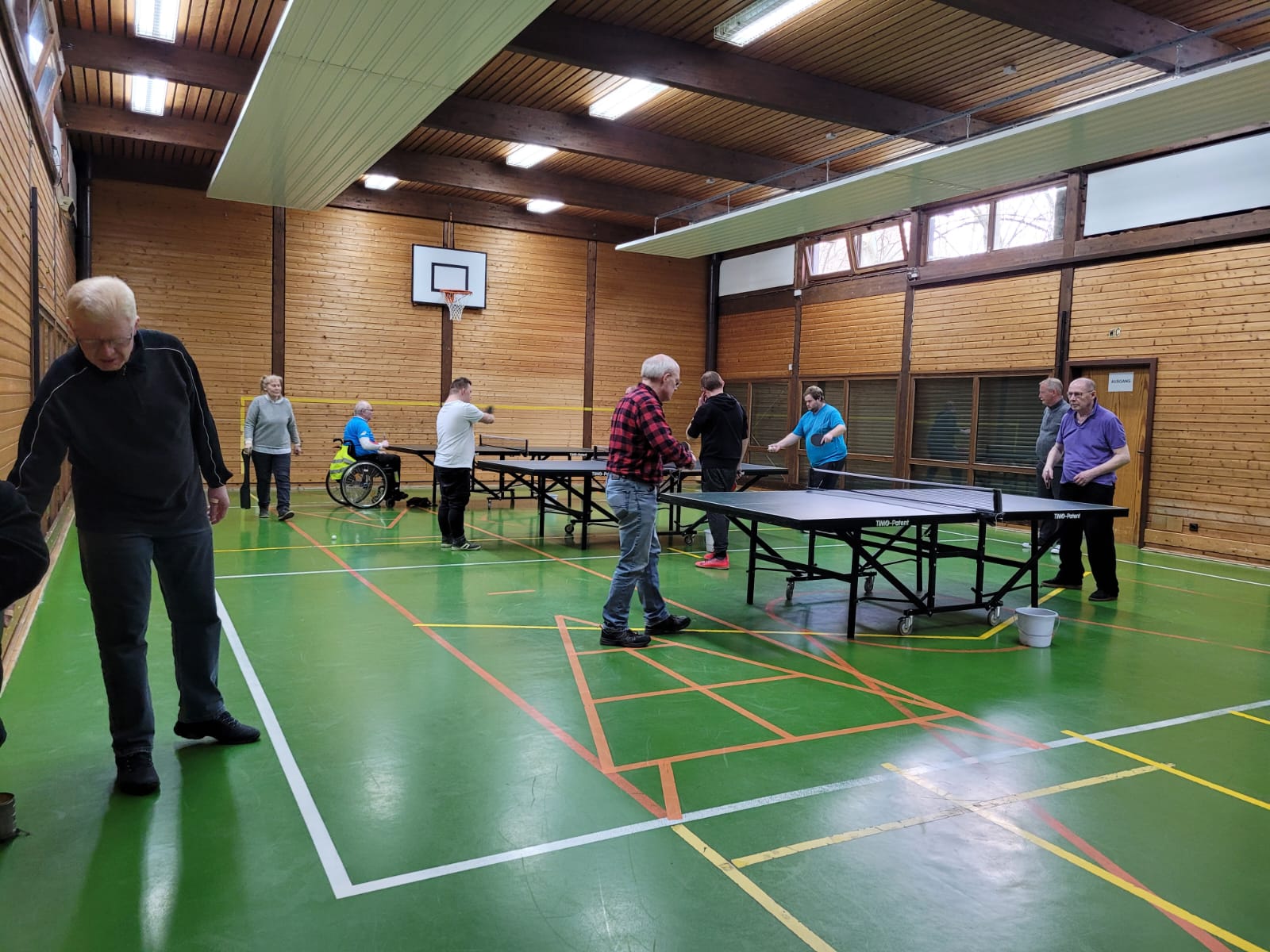 Mehrere Personen spielen Tischtennis in einer Sporthalle, eine Person im Rollstuhl ist ebenfalls beteiligt
