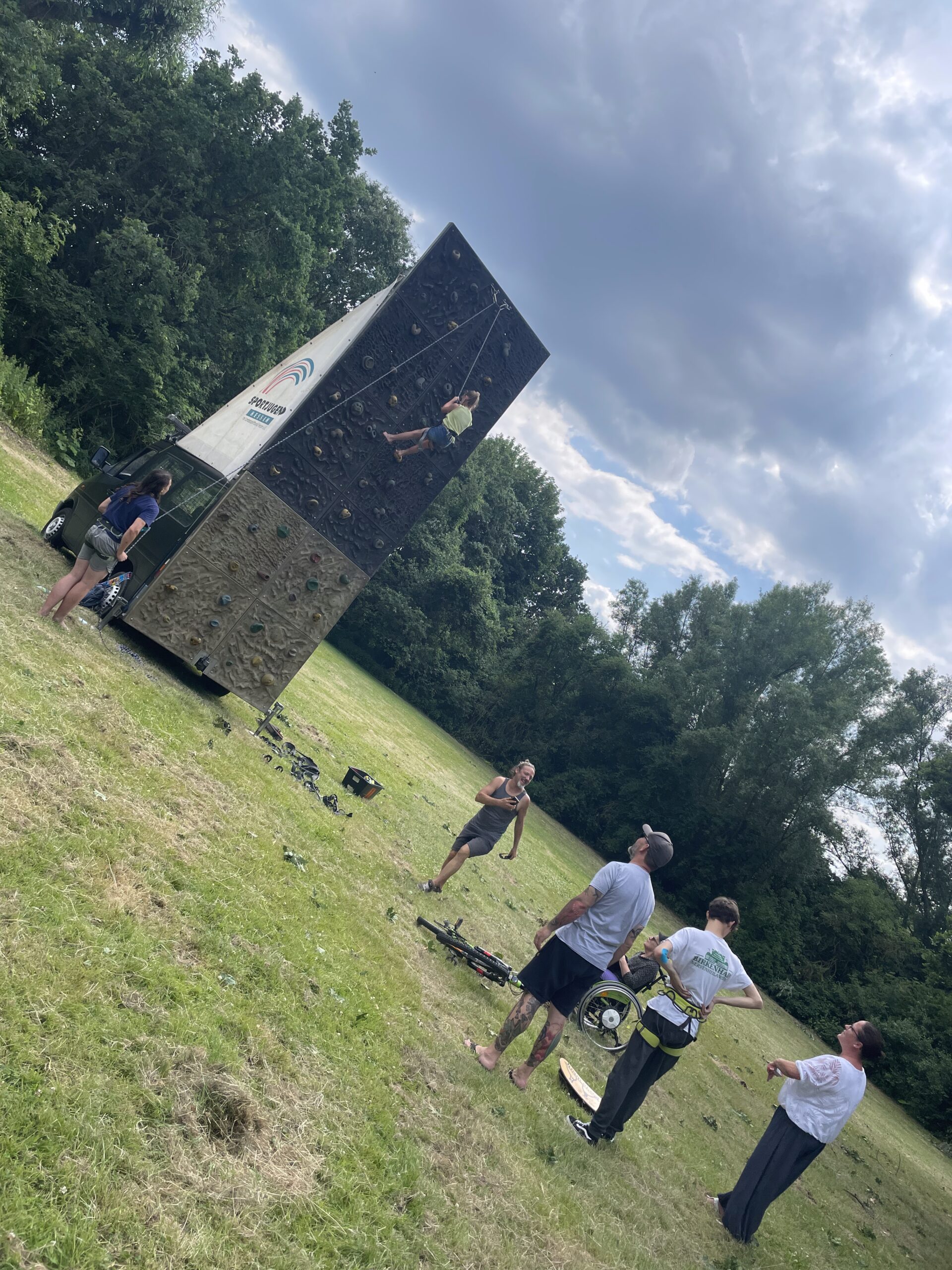 Person klettert an einer mobilen Kletterwand, während andere auf einer Wiese zuschauen. Im Hintergrund Bäume und ein bewölkter Himmel