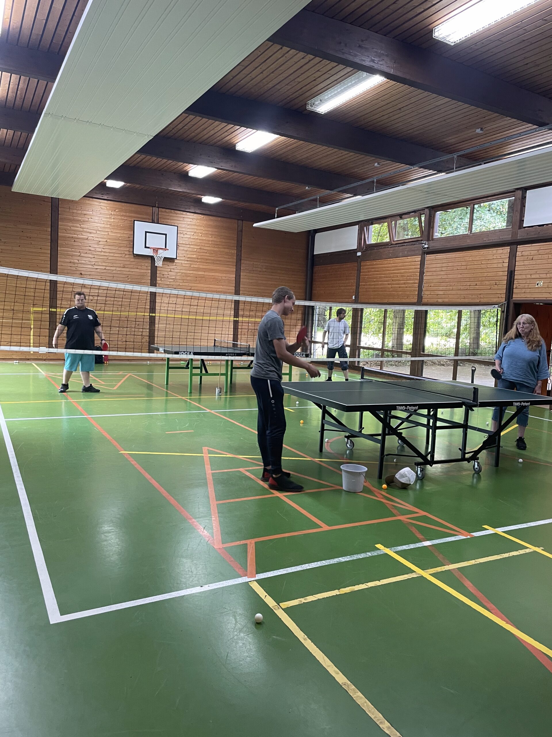 Menschen spielen Tischtennis in einer Sporthalle mit grünem Boden und Holzverkleidung