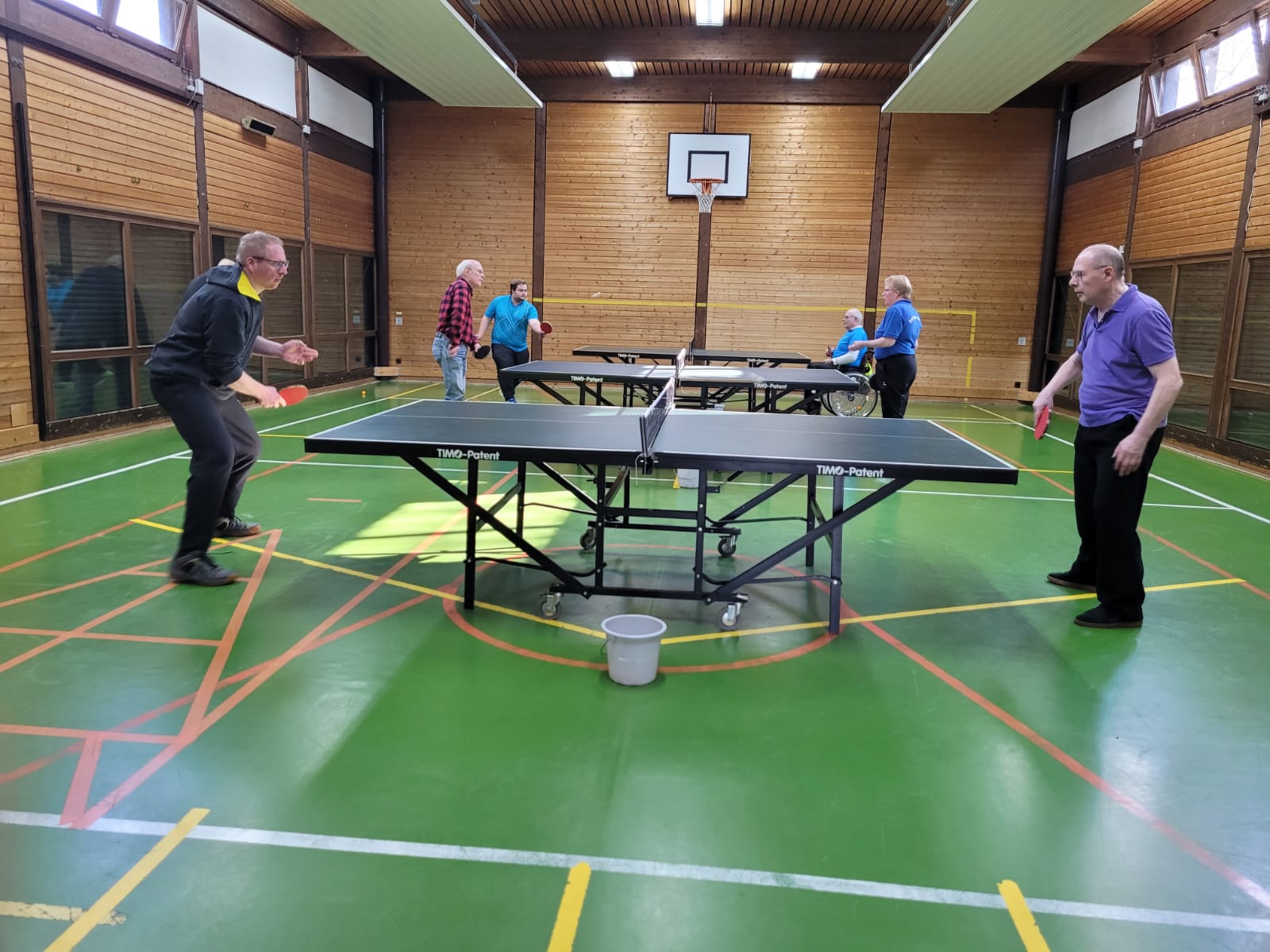 Personen spielen Tischtennis in einer Turnhalle, einige Teilnehmer beobachten oder sind in Rollstühlen.