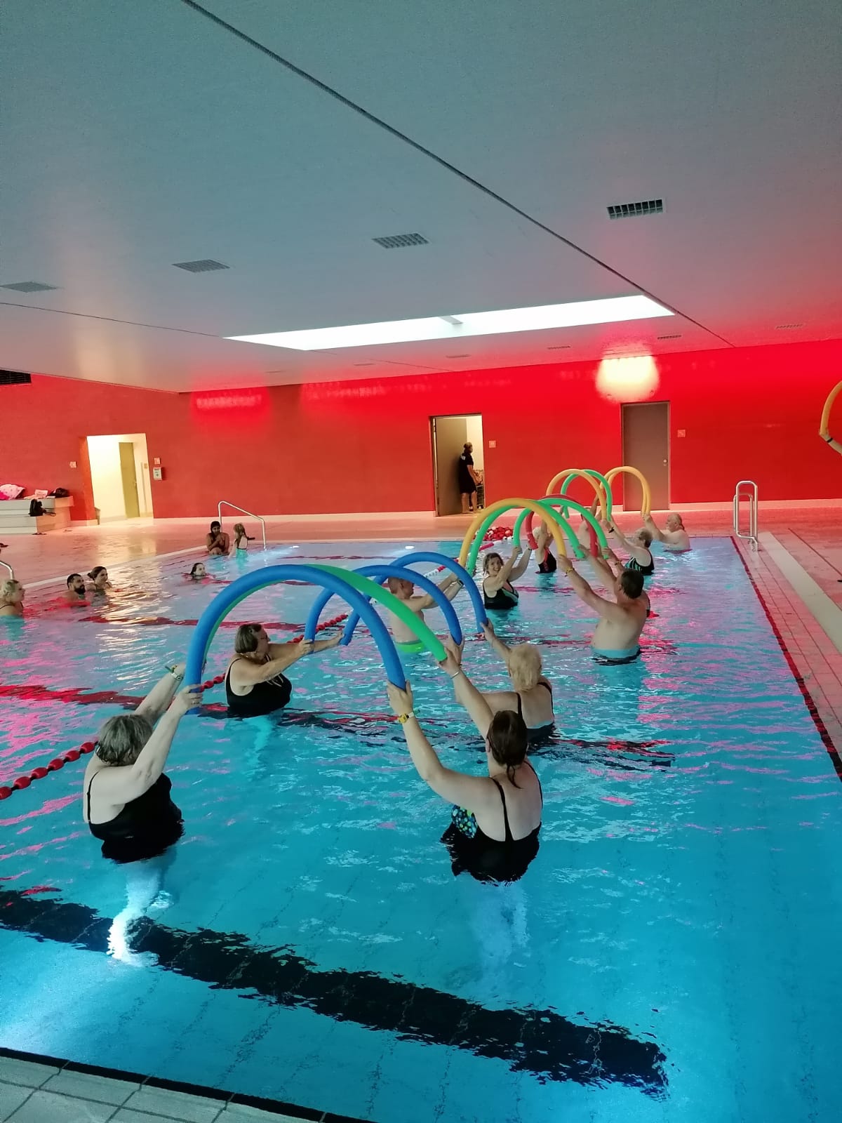 Teilnehmerinnen eines Wassergymnastikkurses in einem Hallenbad, die mit bunten Poolnudeln Übungen durchführen