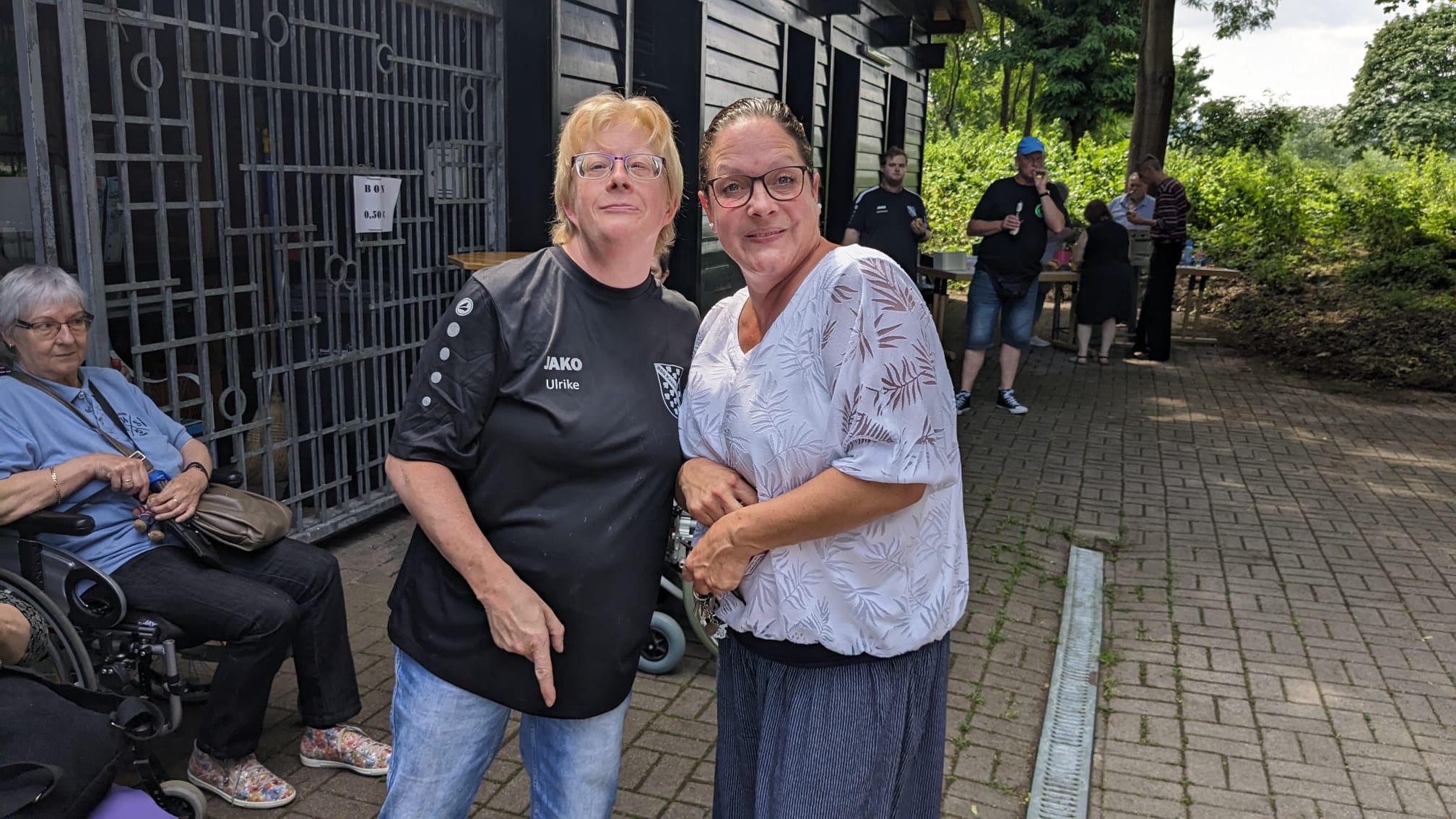 Zwei Frauen lächeln in die Kamera, eine Person im Rollstuhl sitzt im Hintergrund, weitere Menschen sind bei einem Outdoor-Treffen zu sehen