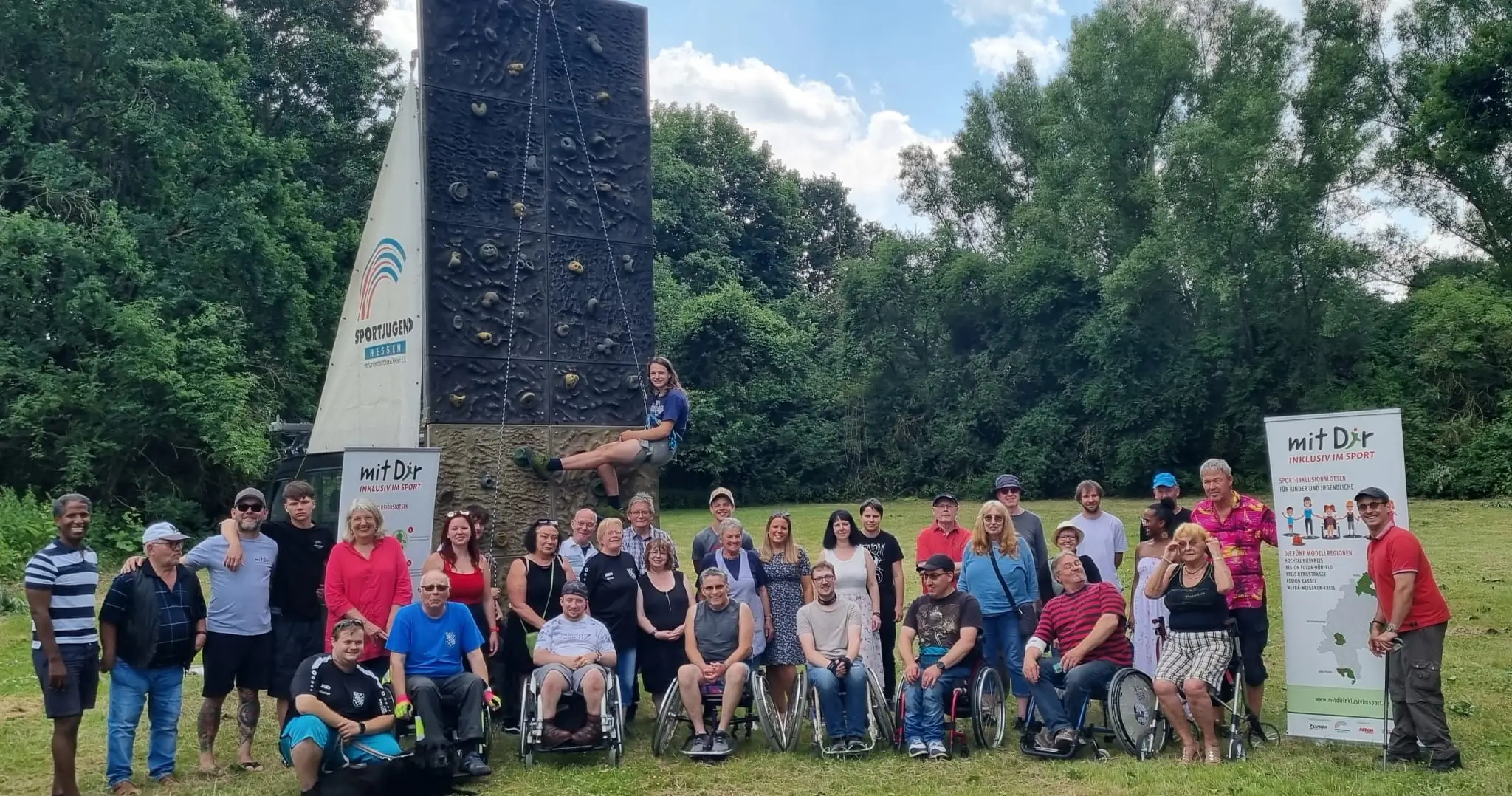 Gruppe von Menschen, darunter Personen im Rollstuhl, posiert vor einer Kletterwand im Freien. Neben der Kletterwand stehen zwei Schilder, eines mit der Aufschrift 'Sportjugend Hessen' und ein weiteres mit 'mit Dir – Inklusiv im Sport'. Im Hintergrund ist eine grüne Waldlandschaft und ein sonniger Himmel zu sehen.