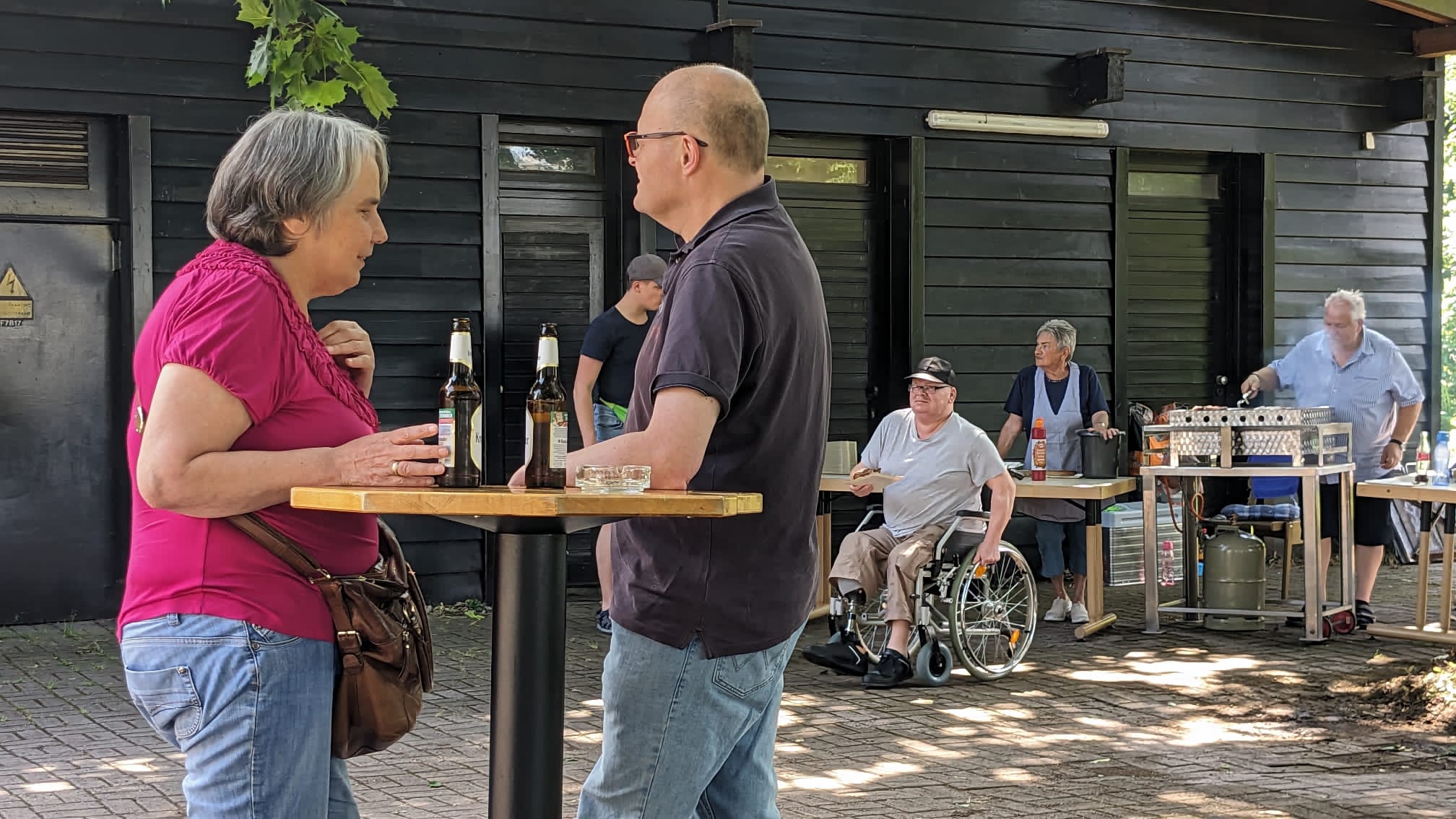 Menschen bei einem Outdoor-Treffen, zwei unterhalten sich an einem Stehtisch, andere sitzen oder bedienen einen Grillstand